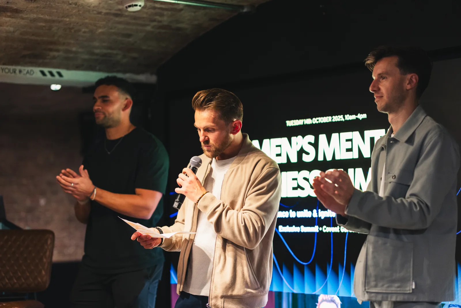 Three men on stage at the Men's Mental Fitness Summit, one speaking into a microphone