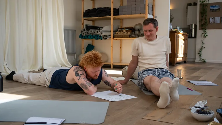 Two men on the studio floor during facilitator training, one sketching a model on paper while the other watches